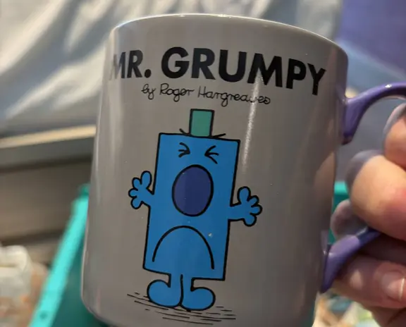 A gray mug featuring Mr. Grumpy cartoon character in blue with black outline, text 'MR. GRUMPY' and signature by Roger Hargreaves. The mug is being held by a person.