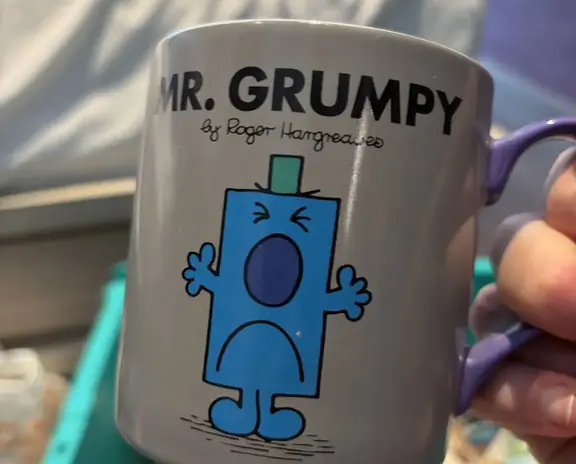 A gray mug featuring Mr. Grumpy cartoon character in blue with black outline, text 'MR. GRUMPY' and signature by Roger Hargreaves. The mug is being held by a person.
