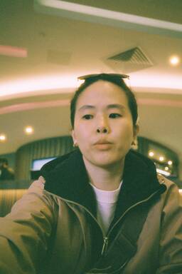 A person taking a selfie in an indoor setting with warm lighting; sunglasses rest on their head and zipper jacket visible.