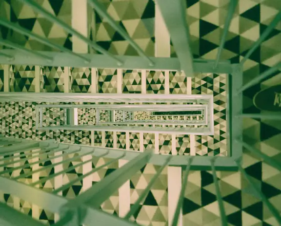 A long, geometric stairwell with repeating triangular patterns on walls and railings, viewed from above downward along the central square staircase.