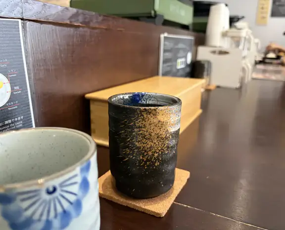 A ceramic cup on a coaster sits on a counter in a cafe; another ceramic cup is partially visible in the foreground. Background shows a menu board and generic counter area.