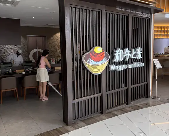 Restaurant storefront with a dark wooden lattice facade and a colorful sign reading "Wagyu and Rice" beside a graphic bowl. Inside, a person stands near the counter.