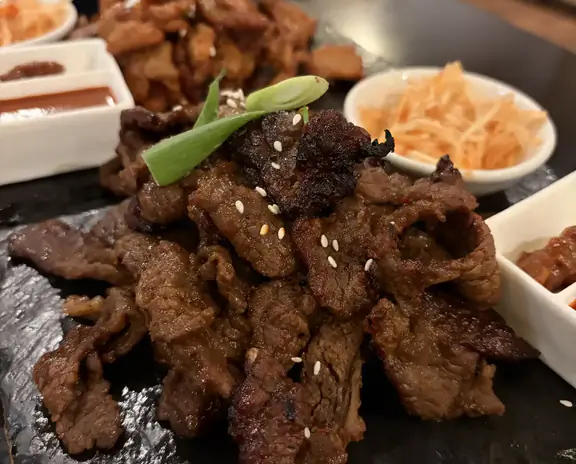 Slices of seared beef arranged on a dark serving board with sesame seeds and green garnish, plus small bowls of sauces and sides in the background.