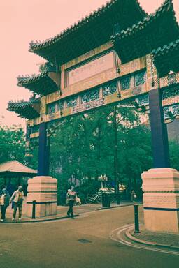 A traditional ornate Chinese arch gate with colorful details, entry path, and people walking nearby in a park setting.
